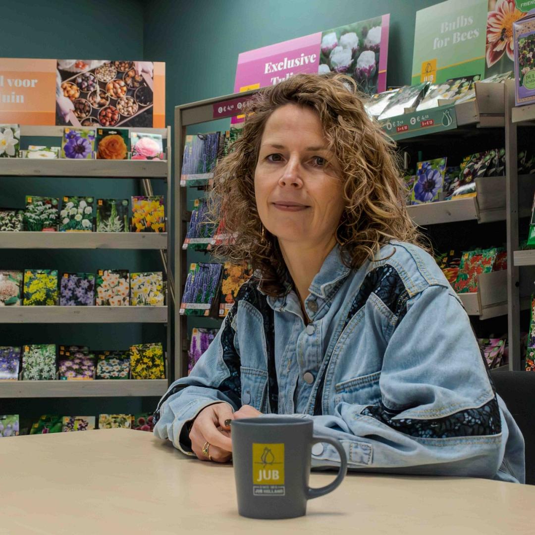 Een vrouw met krullend haar zit aan een tafel met een koffiekopje waarop 'JUB' staat. Ze draagt een spijkerjack met donkerblauwe accenten en kijkt in de camera. Op de achtergrond staan schappen met zakjes bloembollen en kleurrijke verpakkingen.