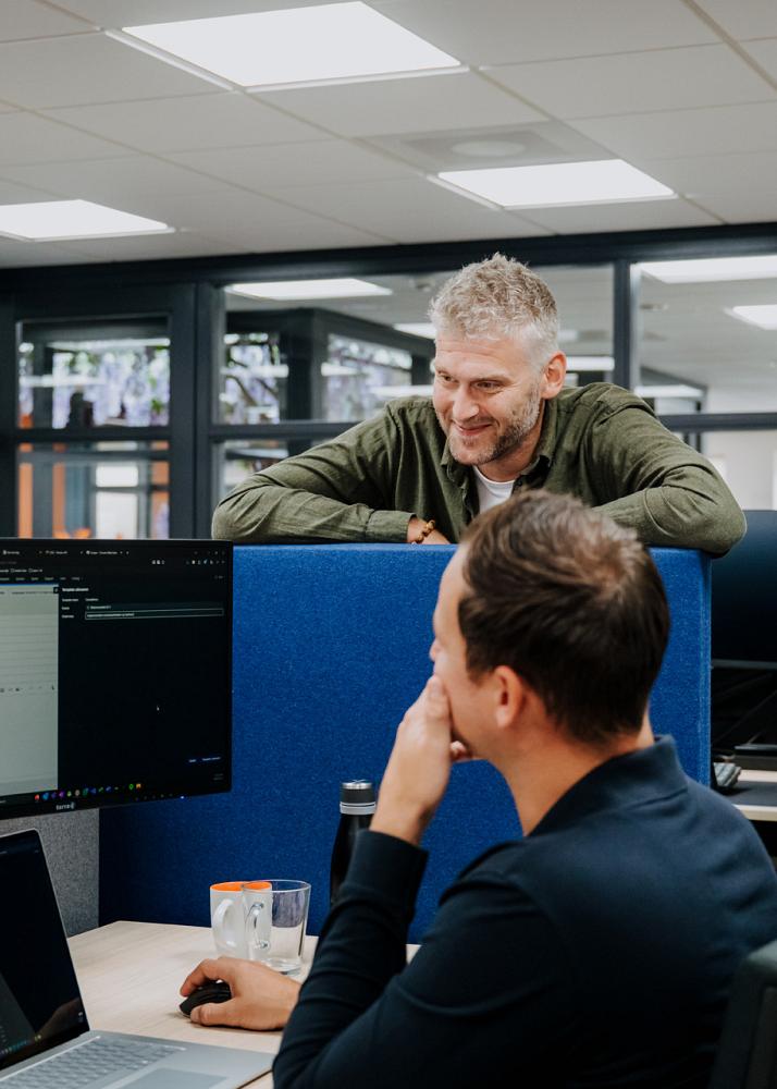 Twee collega’s werken samen achter meerdere computerschermen in een modern kantoor. Eén persoon zit aan het bureau, terwijl de ander lachend meekijkt en overlegt.