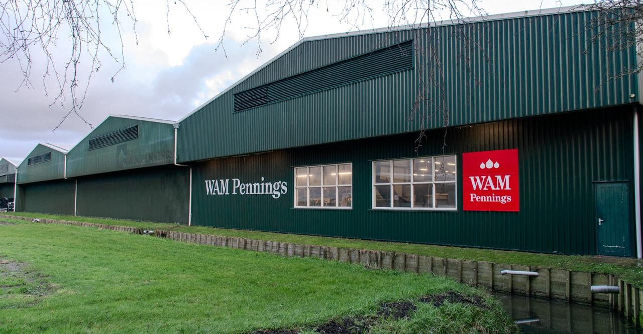 Groen bedrijfspand van WAM Pennings met witte tekst en een rood logo. Voorgrond met gras en een smalle sloot. Kale takken hangen in de bovenhoek.