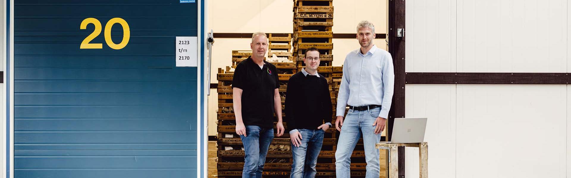 Drie mannen staan voor een open koelcel met het nummer 20, waarin houten pallets zijn opgeslagen. Rechts staat een eenvoudige tafel met een laptop erop.