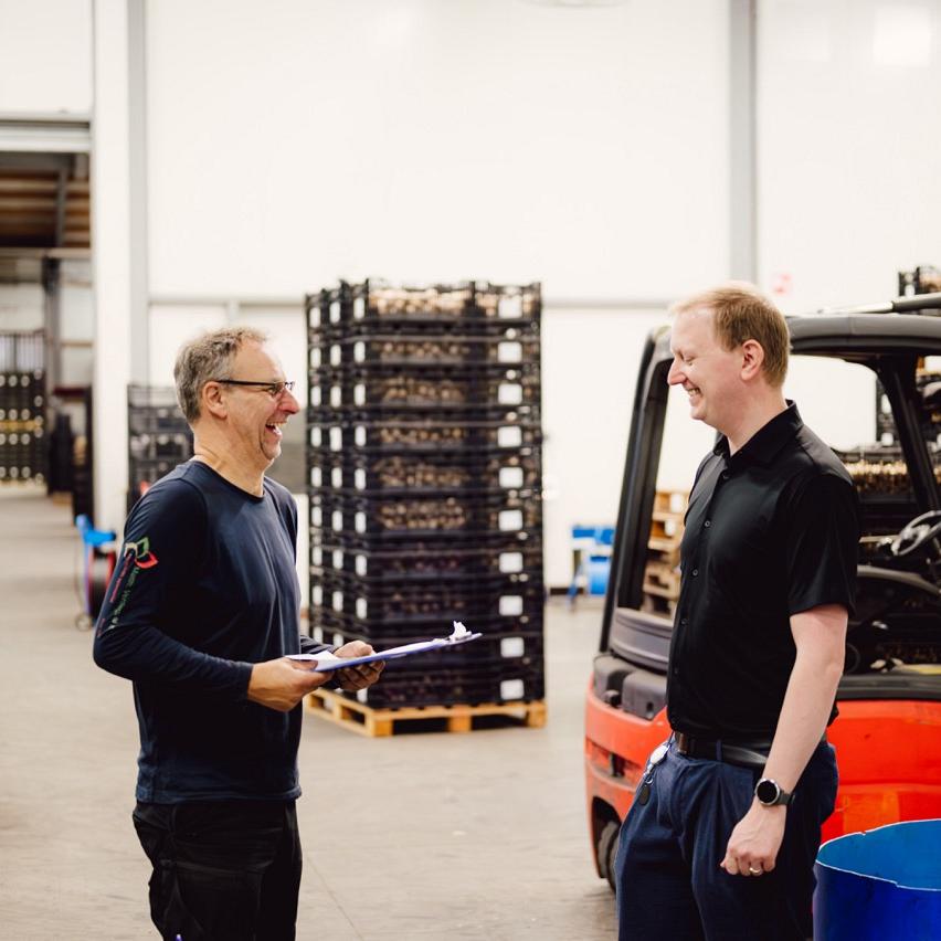 Twee mannen lachen en praten in een magazijn, omringd door stapels kratten en rode heftrucks. Links een medewerker op een heftruck.