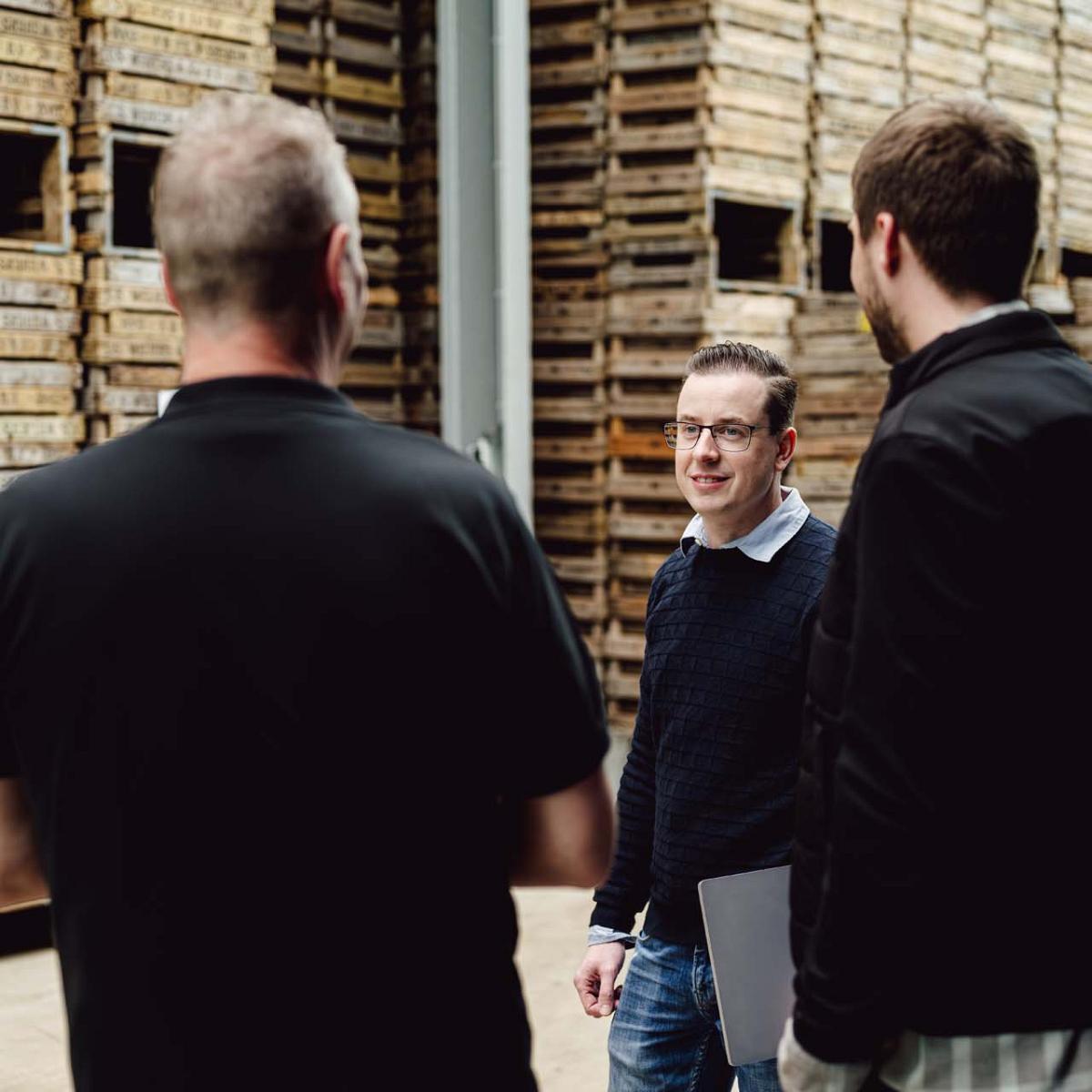 Drie mannen in gesprek in een hal van een bloembollenbedrijf, omringd door hoge stapels houten kisten met bloembollen.