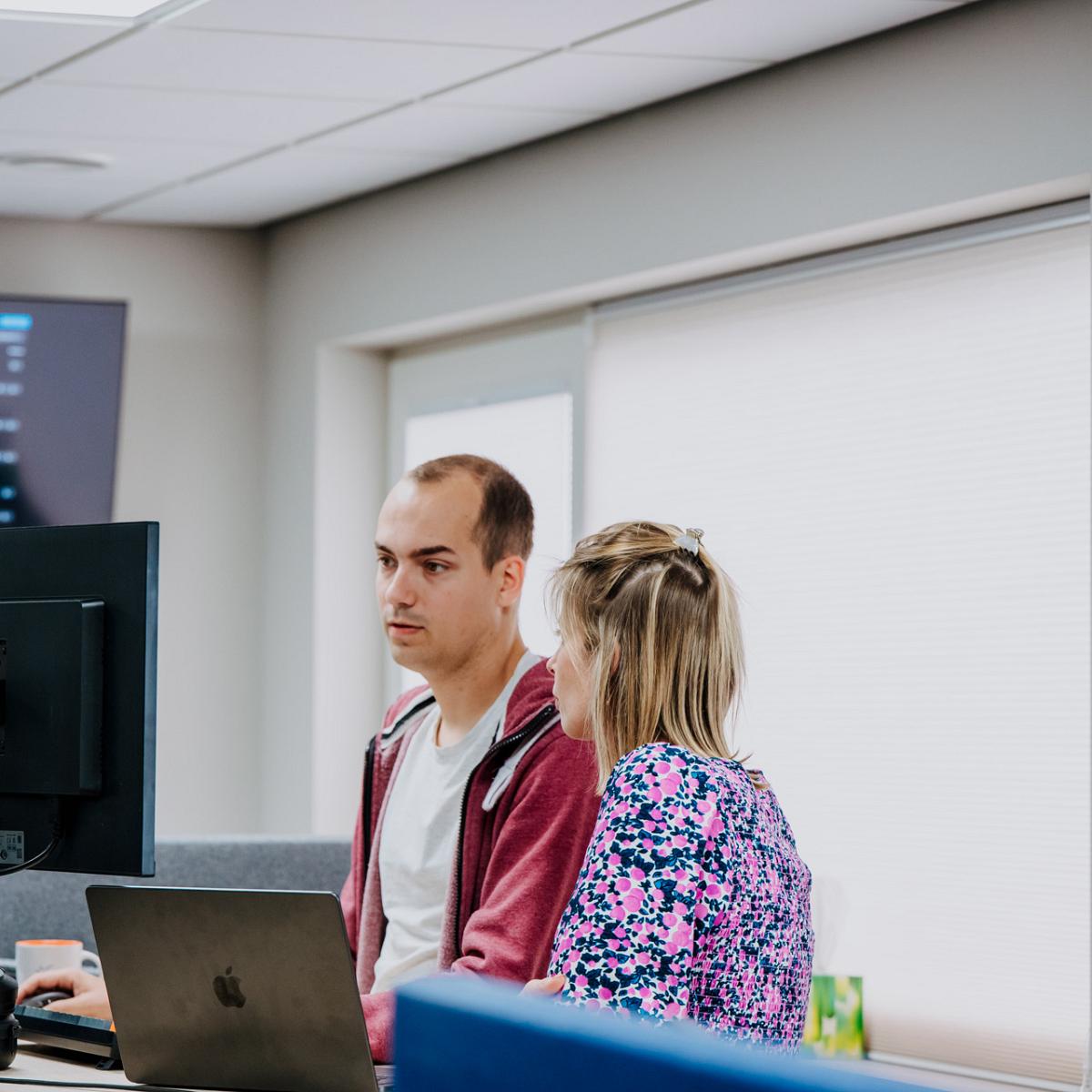 Twee collega’s staan bij een werkplek met computerschermen en een laptop in een kantoorruimte. Ze kijken geconcentreerd naar het scherm terwijl ze overleggen.