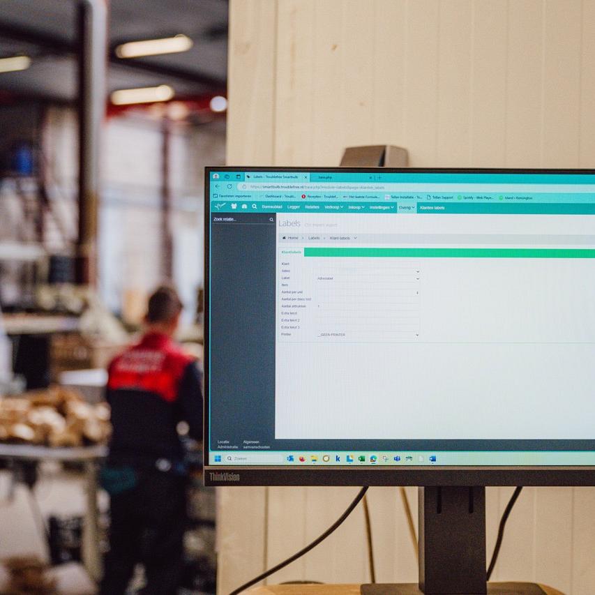 Computerscherm met software in een magazijnomgeving, met op de achtergrond een medewerker en opgeslagen materialen.