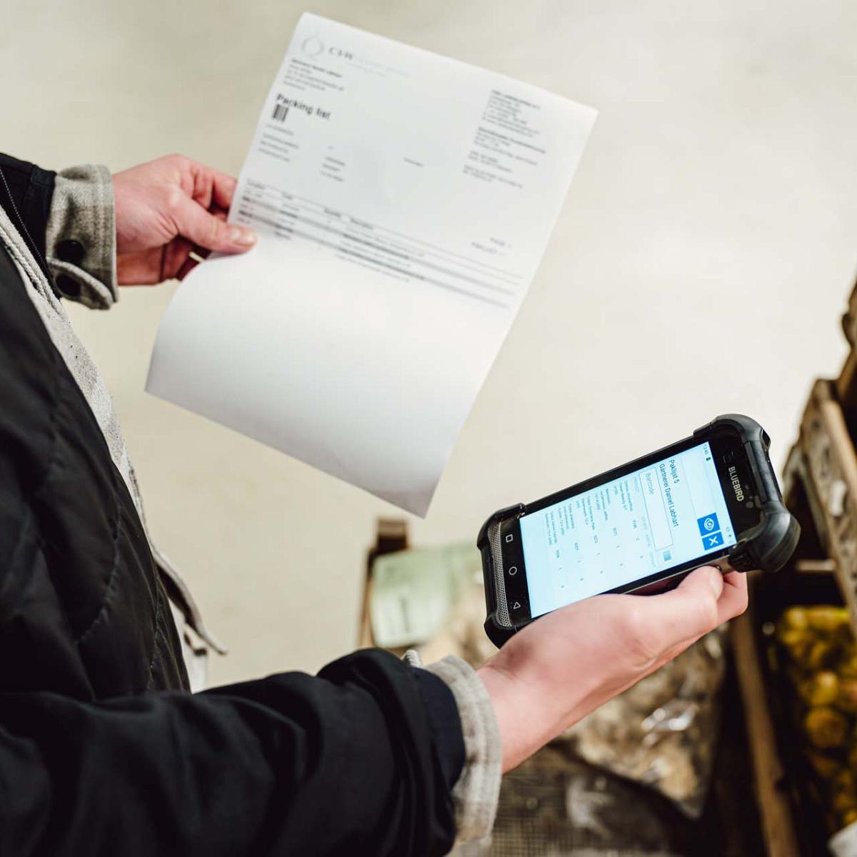 Medewerker controleert een paklijst in de ene hand en een handheld apparaat met gegevens in de andere. Op de achtergrond houten kratten met verpakte producten.