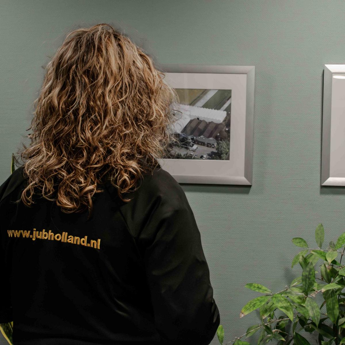 Een persoon met krullend blond haar en een zwarte jas met de tekst 'www.jubholland.nl' kijkt naar twee ingelijste luchtfoto's van landbouw- en bedrijfsterreinen aan de muur.