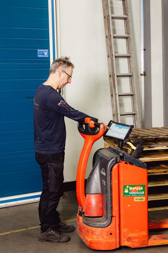 Medewerker bedient een rode elektrische palletwagen met een scherm, staand naast een blauwe magazijndeur met het nummer 10. Op de achtergrond pallets en gestapelde kratten.