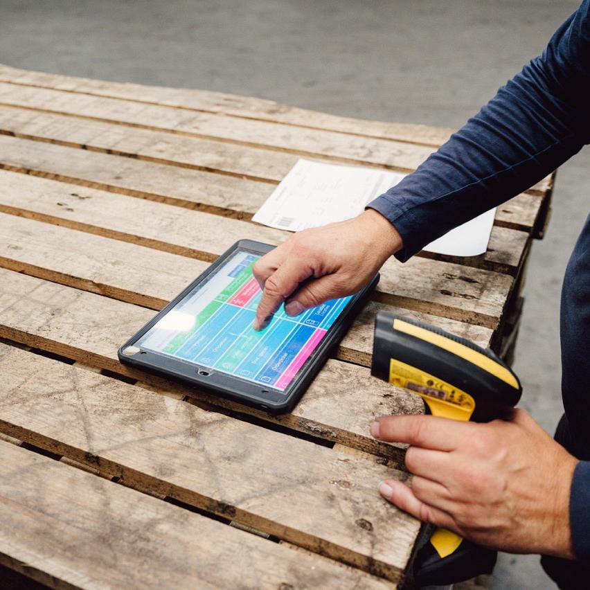 Handen bedienen een tablet met digitale informatie op een houten pallet, naast een handscanner in een magazijnomgeving.