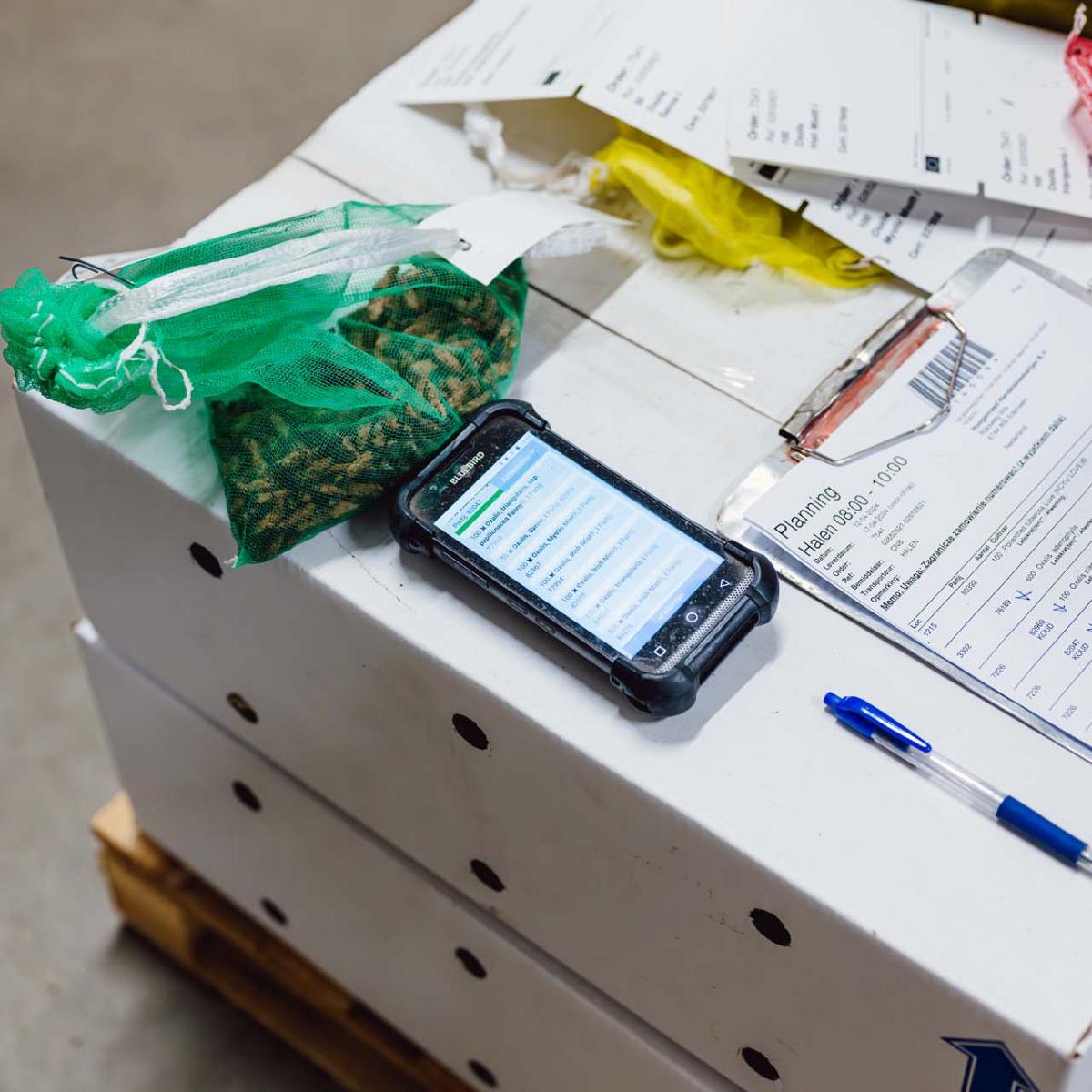 Smartphone naast een klembord met papieren en pen op witte doos naast groene netzak.