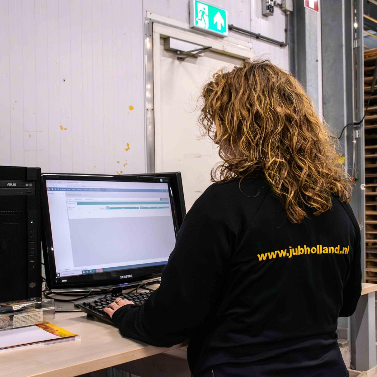 Een persoon met krullend blond haar en een zwarte jas met het logo 'www.jubholland.nl' zit aan een bureau achter een computer, waar het Smart Bulb software op staat.