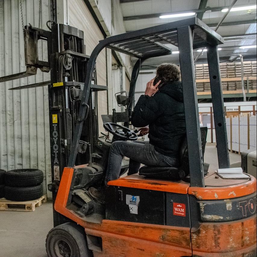 Man in een heftruck van Toyota, bedienend naast een stapel grote houten kratten. Op de achtergrond stapels banden en een magazijn met metalen wanden.
